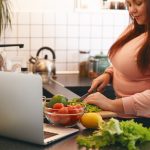 Overweight woman using laptop to watch video recipe while making vegan vitamin avocado salad, slicing leaf lettuce on wooden cutting board. Healthy food, weight loss, dieting and nutrition concept
